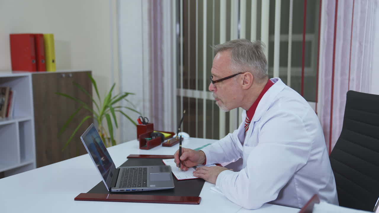 Doctor listens to a patient through the remote webcam on laptop screen. Professional male doctor in hospital office talking to a client online by video call on computer. Profile view.