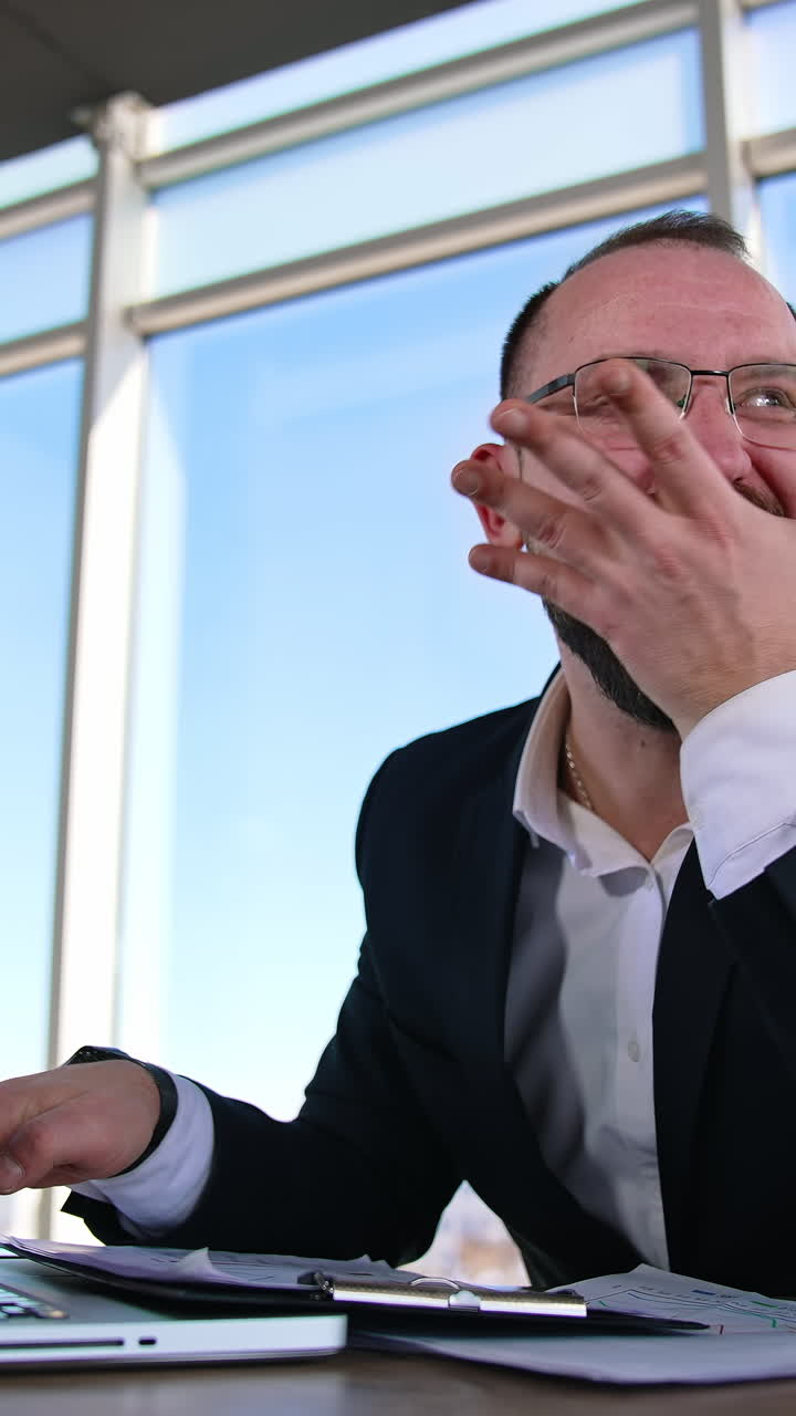 Funny businessman typing on a laptop. Office worker in glasses sitting at his desk in workplace and making mad face. Problems in business. Vertical video