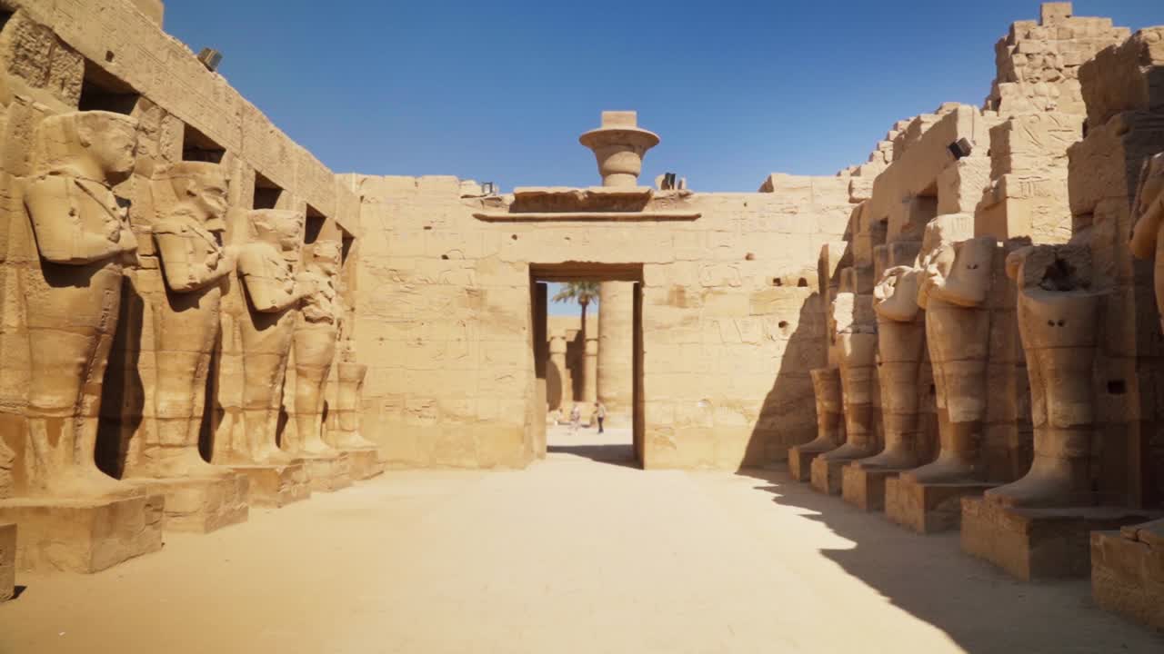 An entry way into the Kamak Temple with tall statues lining each side on a sunny day in Luxtor, Egypt. Static shot with a person in the distance to show the massive scale.