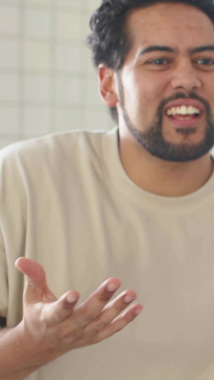 Vertical video: Facing camera, man explaining and gesturing indoors by tiled wall in beige T-shirt
