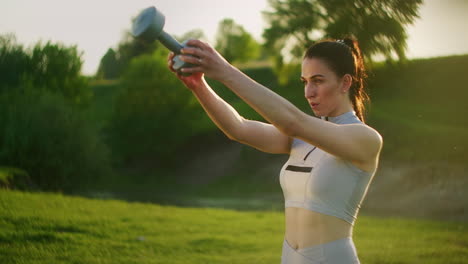 una mujer deportiva en un parque en cuclillas con pesas en las manos al atardecer. entrenamiento con pesas al aire libre. fitness en un parque público en verano para los muslos y las piernas.