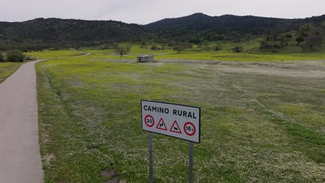 Vuelo-Con-Dron-Visualizando-Un-Prado-Lleno-De-Flores-Blancas-Y-Amarillas-Con-Un-Cartel-Indicando-Un-Camino-Rural-Y-Su-Normativa,-Dicho-Camino-Esta-A-Un-Lado-Vemos-Un-Fondo-De-Montañas-Abulenses