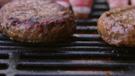 burgers being gently cooked by fire flames hot bbq with pork belly slices in background. close up slow motion barbecue summer cooking food.