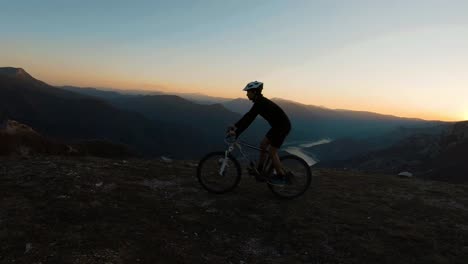 Hombre-Montando-En-Bicicleta-En-La-Cima-De-Una-Montaña-Con-Un-Hermoso-Lago-Del-Cañón-Al-Atardecer-En-El-Fondo