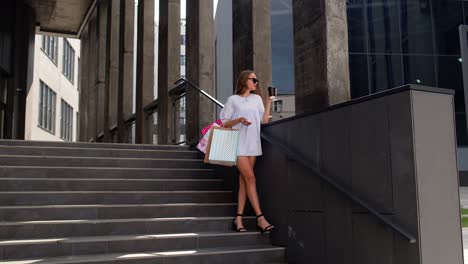 girl with phone staying on shopping mall stairs with colorful shopping bags and drinking coffee