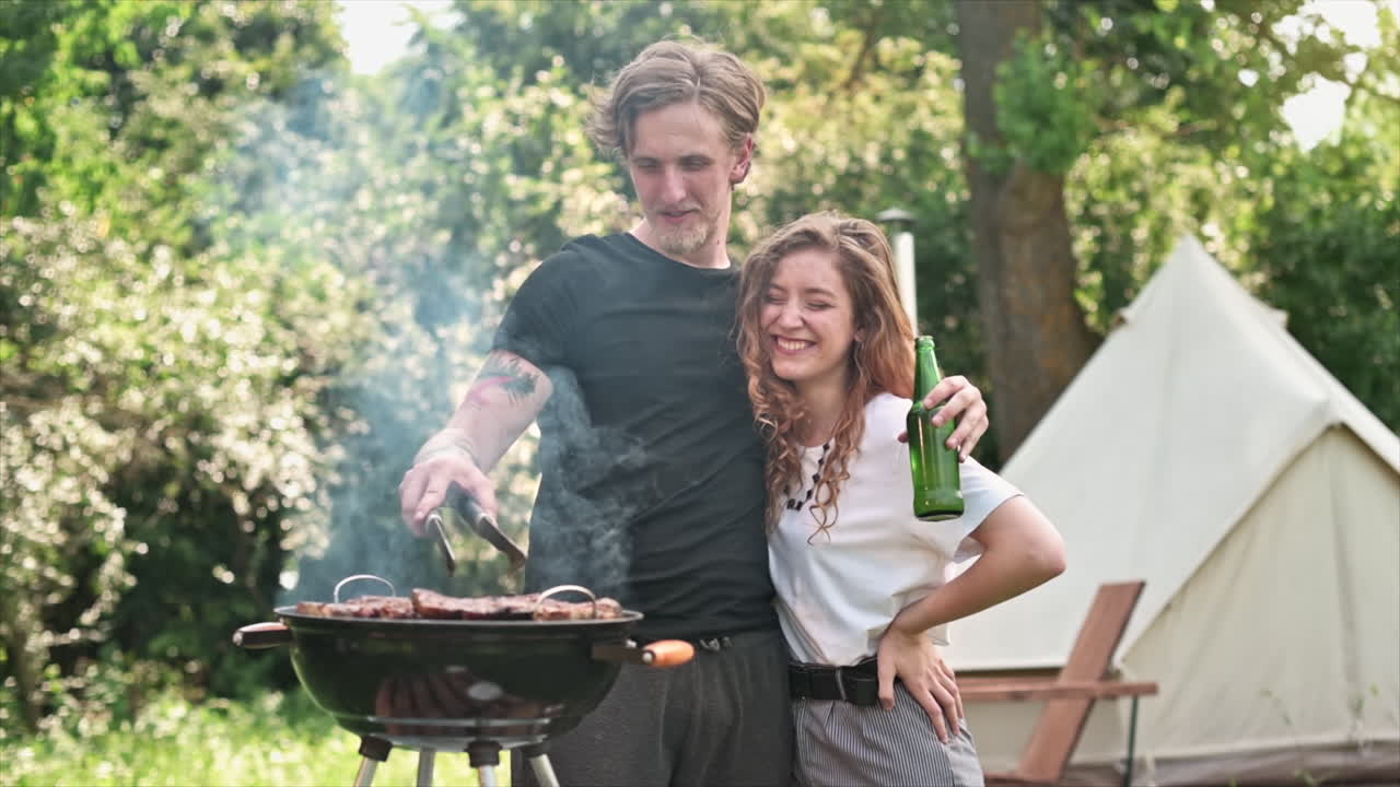 Young couple frying meat on the grill and drinking beer, laughing. Greenery around. Glamping. Slow motion