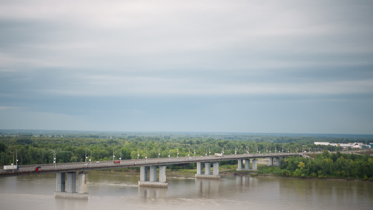 흐린 하늘 아래 넓은 강 다리, 긴 콘크리트 경간이 나무가 늘어선 수평선과 부드러운 빛, 잔잔한 물결이 있는 흙탕물을 가로지르며 텅 빈 도로가 통근 정체를 연상시키는 항공 측량 및 엔지니어링에 이상적입니다.