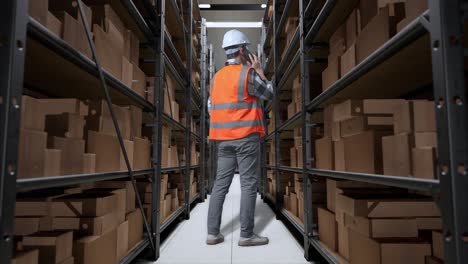 vista trasera completa del cuerpo de un ingeniero masculino asiático con casco de seguridad hablando en teléfono inteligente mientras está de pie en el almacén con estantes llenos de productos de entrega
