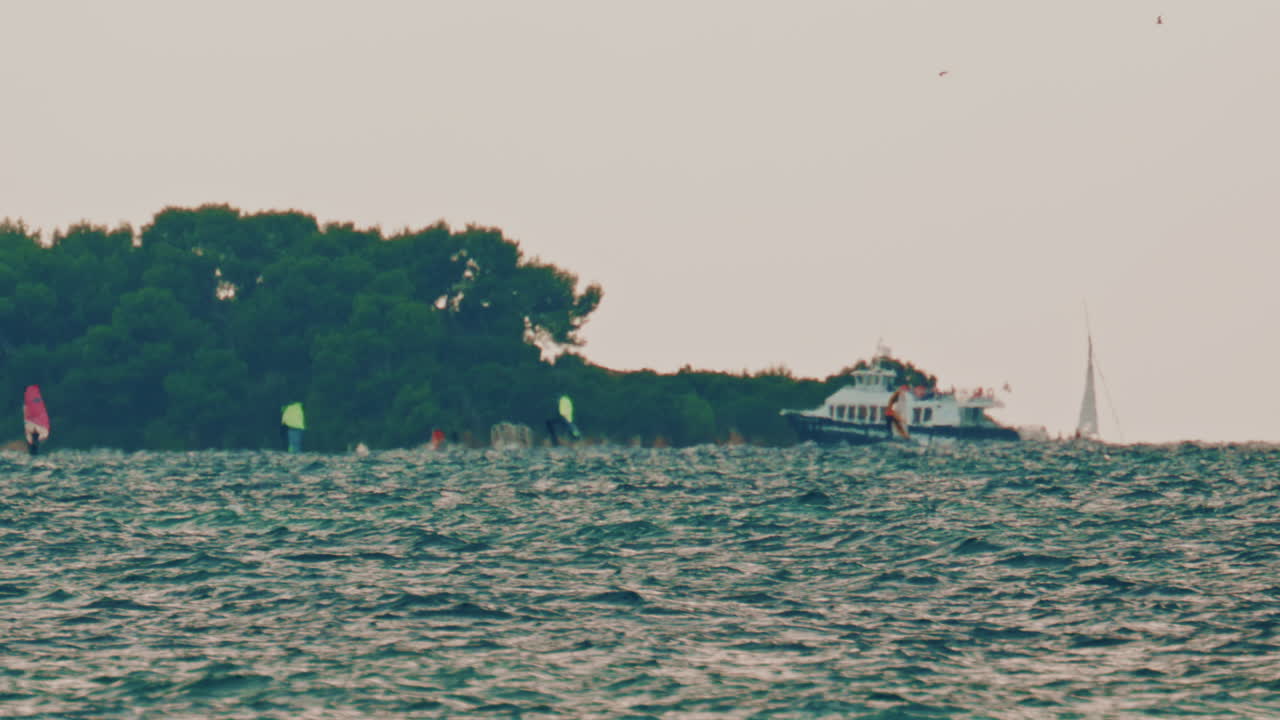 Colourful windsurfers and a motorboat moving along a choppy sea in front of a small wooded island on the French Riviera
