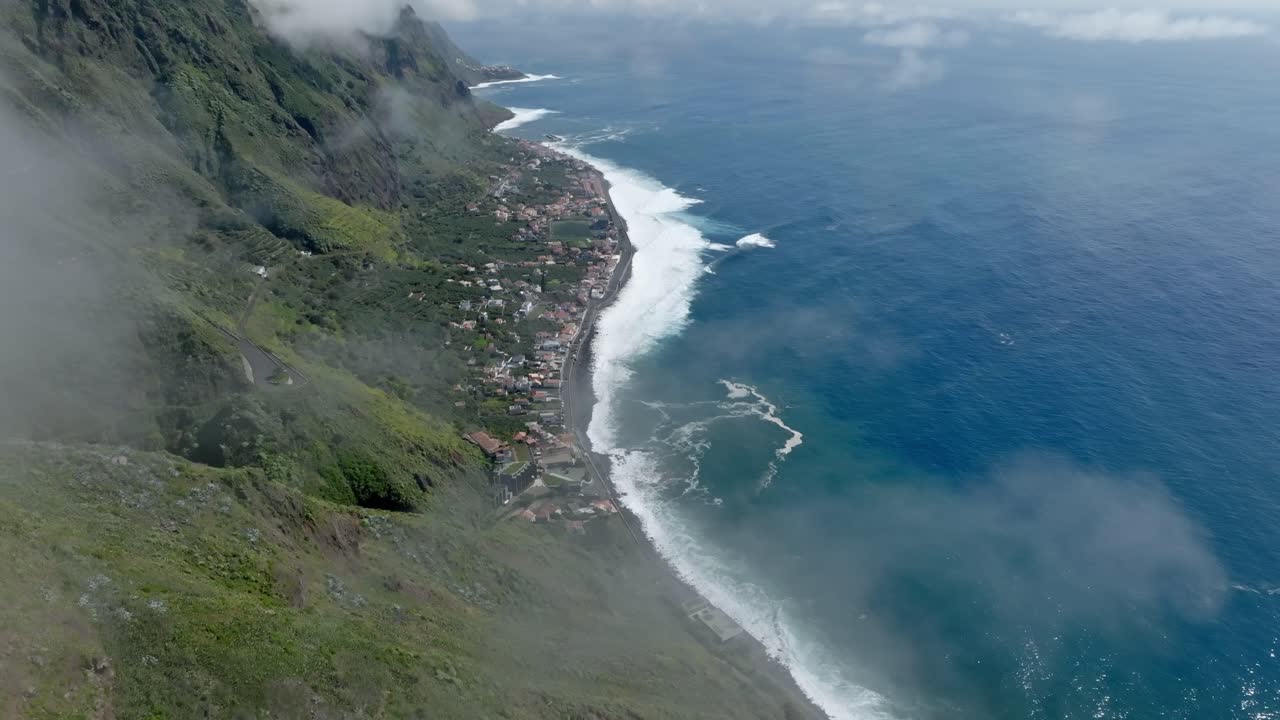 Nubes finas y tenues cuelgan bajas sobre montañas verdes que se elevan sobre un pueblo costero en Madeira, Portugal.