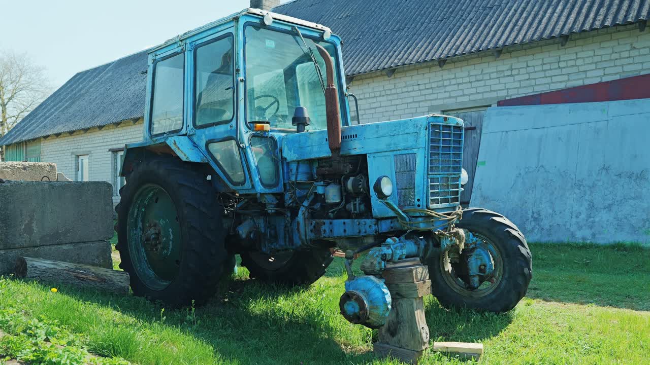 Blue Soviet tractor missing wheel in bright sun beside rural farm building