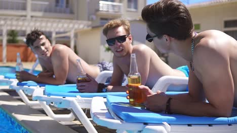 group of young friends drinking beer and relaxing lying on the coaches by the swimming pool