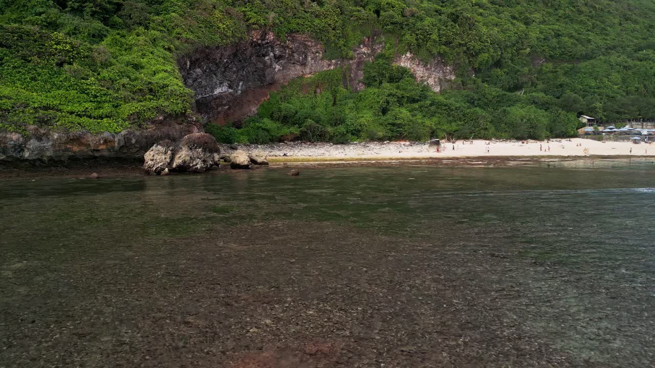 Aerial drone footage capturing Nyang Nyang Beach in Uluwatu, showing travelers walking beneath green cliffs and exploring the rocky shoreline of this peaceful hidden Bali destination