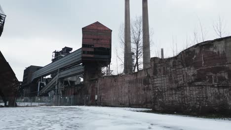 desolate, snow-covered, abandoned industrial site with rusted structures, a tall brick building, and two smokestacks under an overcast sky