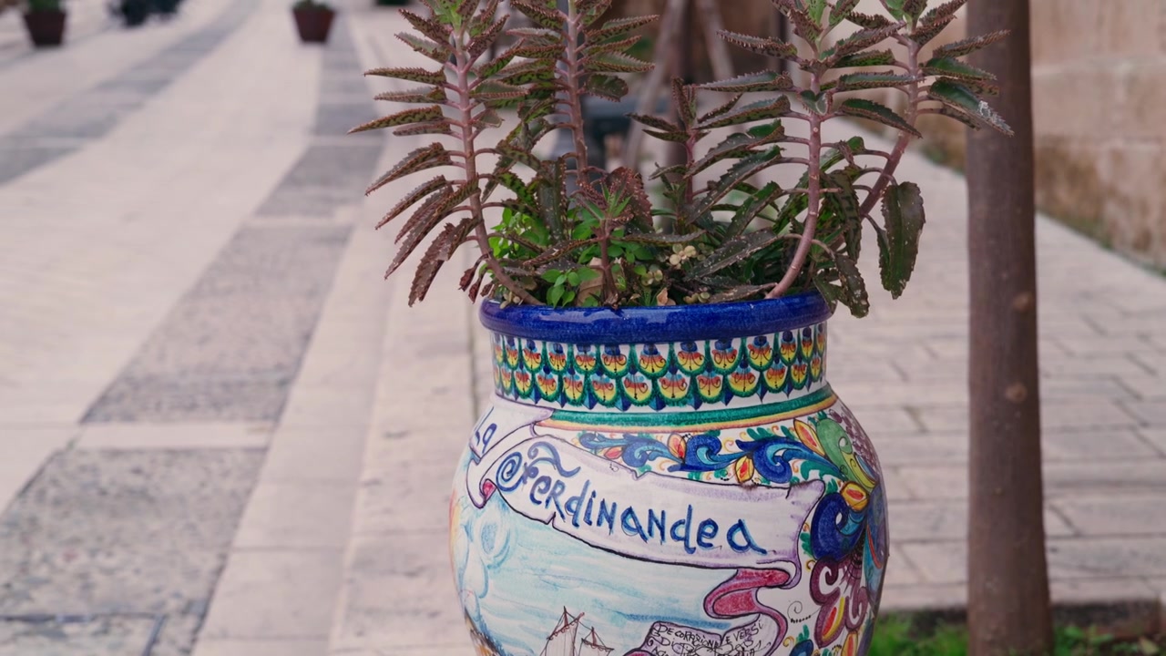 Primo piano di un vivace vaso di fiori in ceramica dipinto a mano, decorato con arte popolare tradizionale siciliana, in una piazza illuminata dal sole.