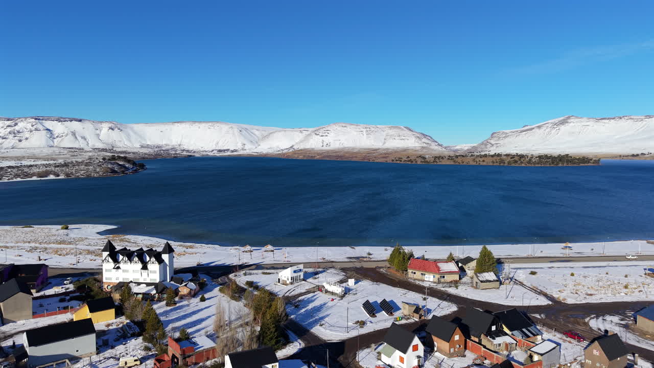 Drone moves backward over snowy buildings near Caviahue lake, showing water, mountains, and winter landscape in Neuquén, Argentina