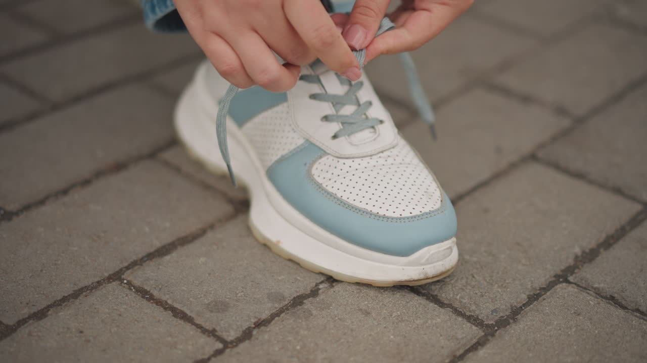 Primo piano di mani che allacciano una sneaker bianca e verde acqua su pavimentazione grigia con risvolto di jeans, calzino nero fantasia, movimento di allacciatura deliberato, atmosfera urbana casual, luce naturale soffusa, pelle traforata testurizzata e spessa.