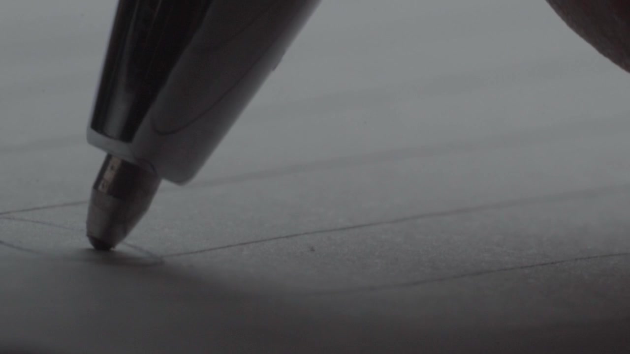 Close-up of a pen writing on a sheet of paper. The writing action is shown in detail, focusing on the pen tip, handwriting, ink marks, and paper surface