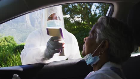 health worker in protective clothes measuring temperature of african american senior woman in car