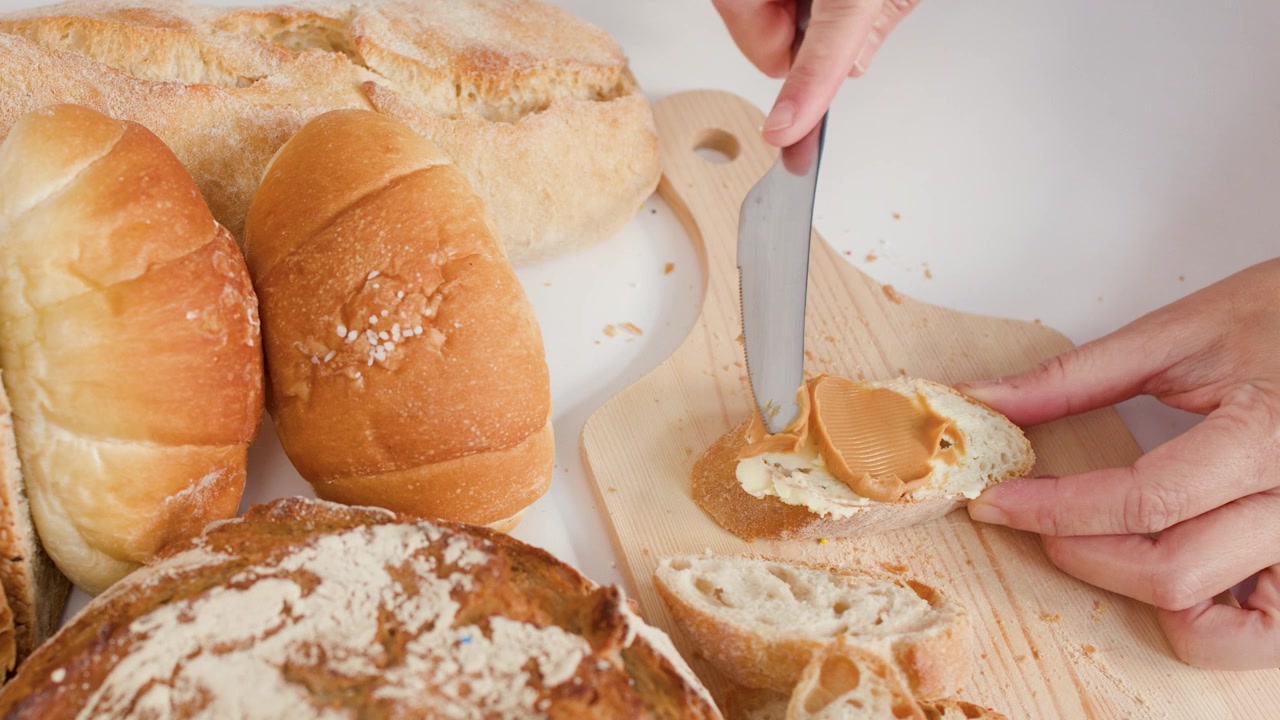 Close-up shot of hands spreading peanut butter on crusty bread amidst various bakery loaves.