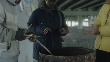 homeless people gather around a barrel for warmth under a bridge