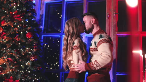 young couple have romantic time near the christmas tree. man and woman in knitted winter sweaters kissing near the new year tree at home.