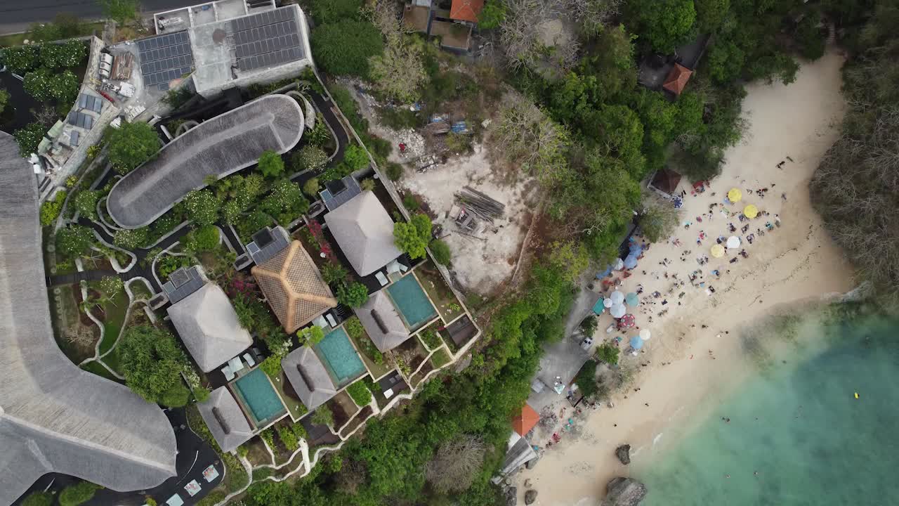 A top-down 4K drone view of a luxury resort and villas nestled against a white sand beach in Uluwatu, Bali. Features multiple private pools, thatched roofs, and turquoise ocean water