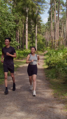 Video-Vertical-De-Una-Pareja-Haciendo-Ejercicio-Al-Aire-Libre-Corriendo-Por-Una-Pista-A-Través-Del-Bosque-Hacia-La-Cámara-Con-Ropa-Deportiva-Filmada-En-Tiempo-Real