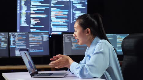 side view of asian female programmer having a meeting by a laptop while writing code on multiple monitors showing database on terminal window desktops in the office