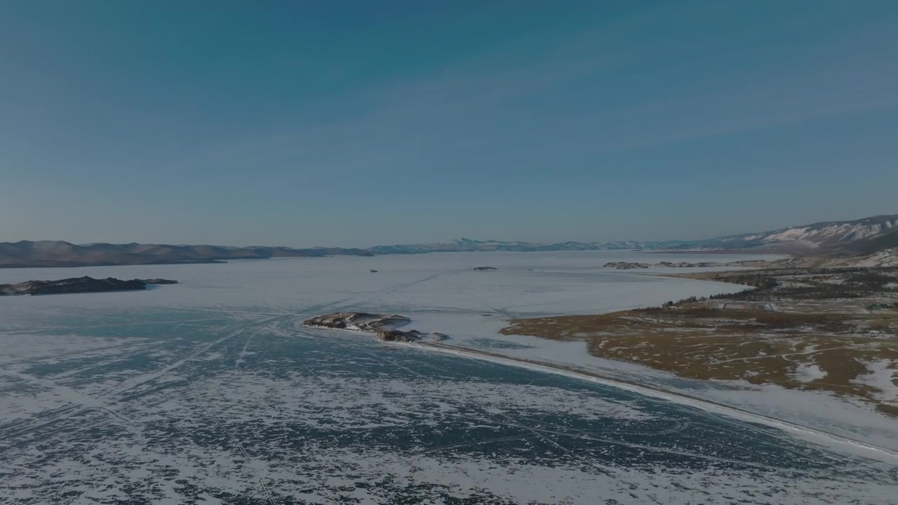 Luftopptak av den enorme frosne Bajkalsjøen med teksturerte ismønstre, små øyer og en fjern fjellkystlinje under en blek vinterhimmel i Russland.