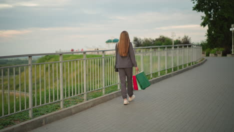 back view of lady in grey clothing walking along iron rail holding shopping bags and a black handbag, with a blurred view of people in the distant background