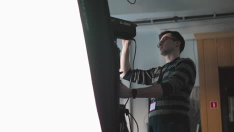 studio technician operating sl-200w light, adjusting position and power settings on light panel with white backdrop, adjusting camera gear for broadcast production scene in studio