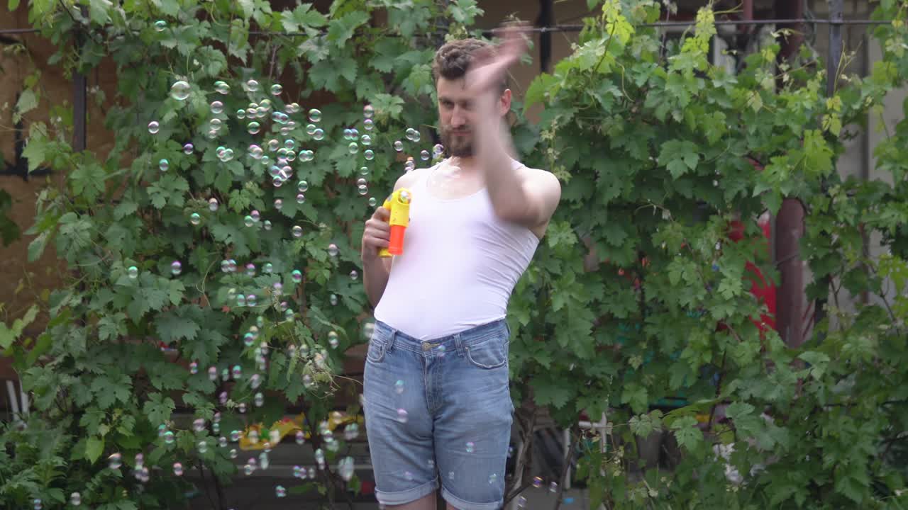 수염을 가진 유아적인 젊은 남자가 서서 장난감 거품에서 비누 거품을 아내고 손으로 흔들고 있습니다.