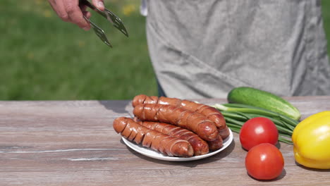 ready grilling sausages with vegetables on the table outdoors. chef puts roast sausage into a plate. tasty meal for a picnic.