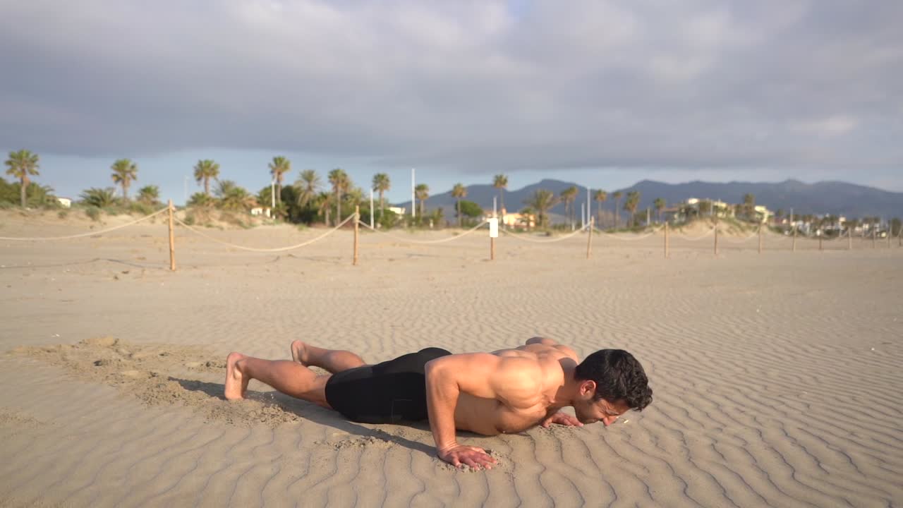 muscular man shirtless performing push-ups variant with jump on beach with palm trees at sunrise