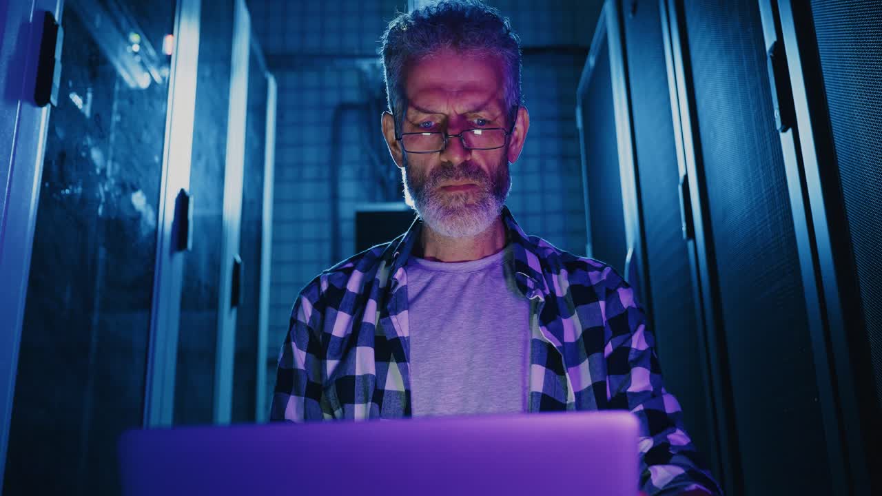 Older Man Working on a Laptop in a Data Center at Night