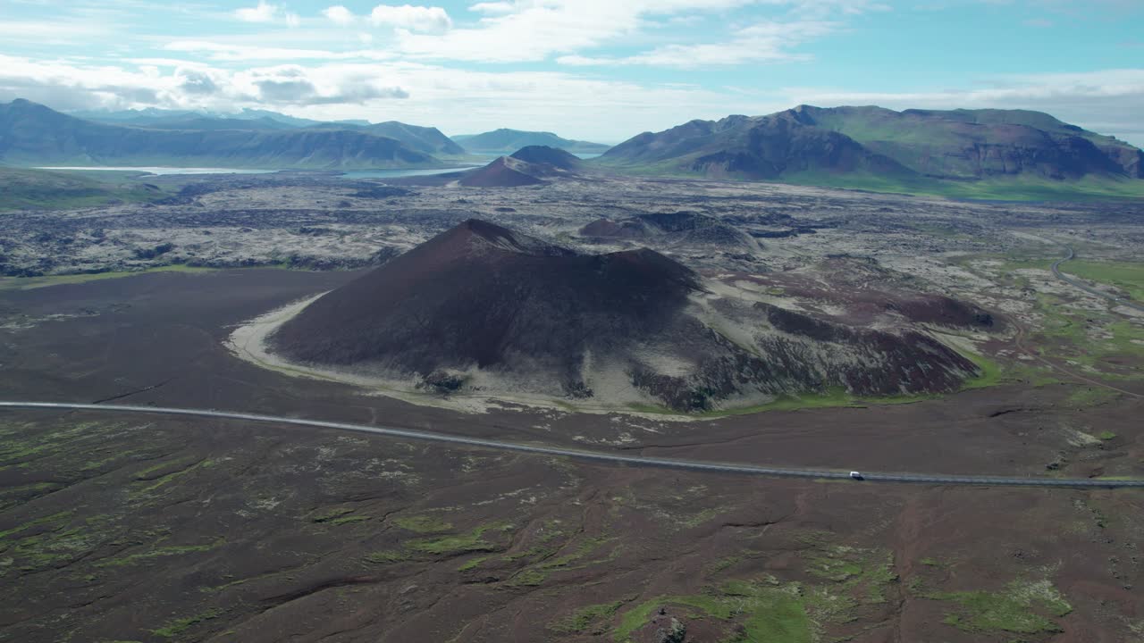An aerial shot of a large volcanic crater mountain located alongside a scenic road in Iceland. The rugged volcanic formation contrasts with the open road winding through Icelandic landscape.
