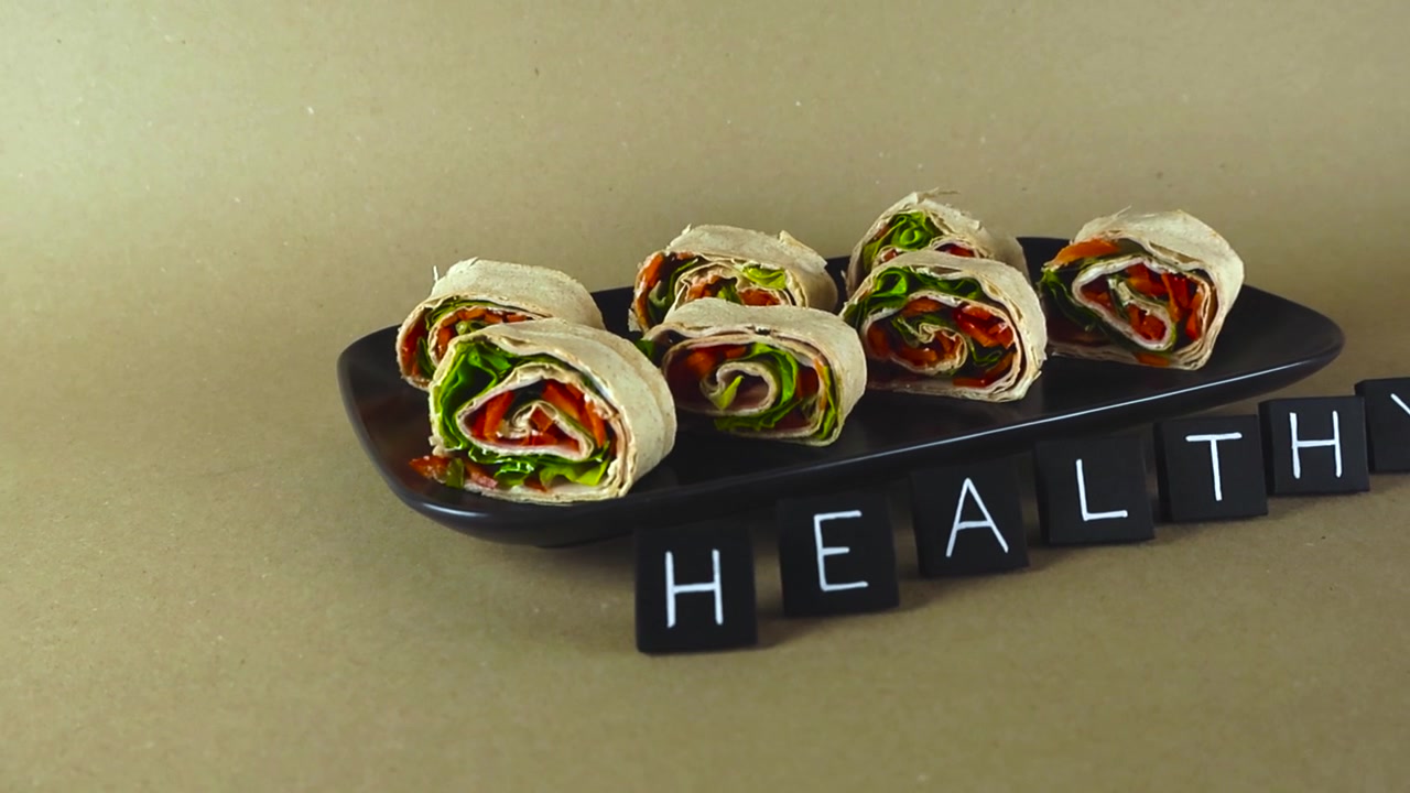 Fresh vegetable and ham lavash pinwheel rolls on a black plate, with black letter tiles spelling the word HEALTHY, representing a wholesome lifestyle, proper nutrition, and mindful eating habits