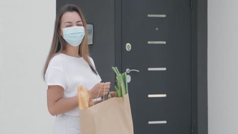 una hermosa mujer con máscara de protección se encuentra con un repartidor que lleva máscara y guantes que le da un paquete de compras con comida al lado de su casa con seguridad. entrega de alimentos, entrega a casa en cuarentena