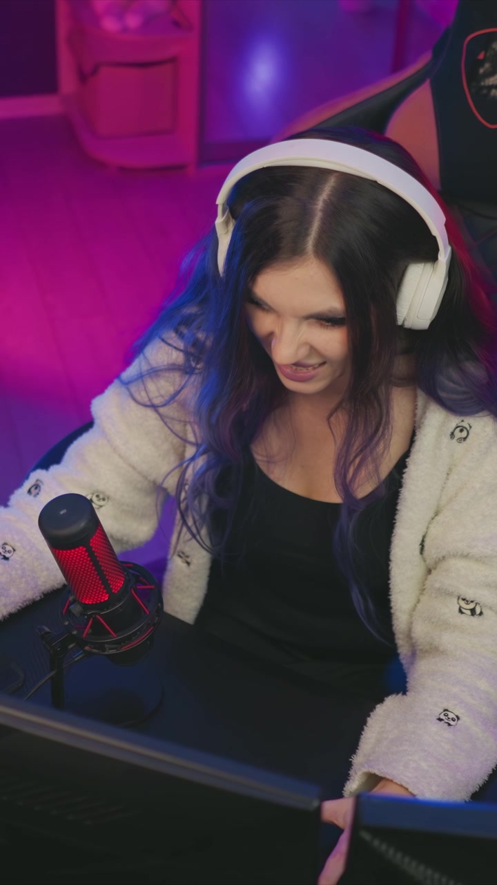 Giovane donna con lunghi capelli scuri seduta su una sedia da gaming, con cuffie bianche e giacca soffice. La donna interagisce in modo discreto con lo schermo del computer, circondata da una luce ambientale rosa