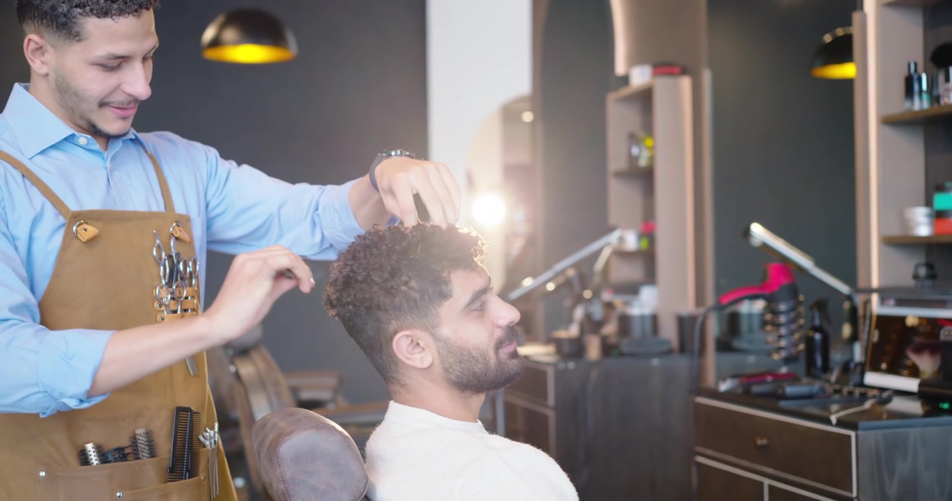 Afroamerikansk mandlig barber og mandlig kunde former krøller med kam og klippemaskine i salonen.