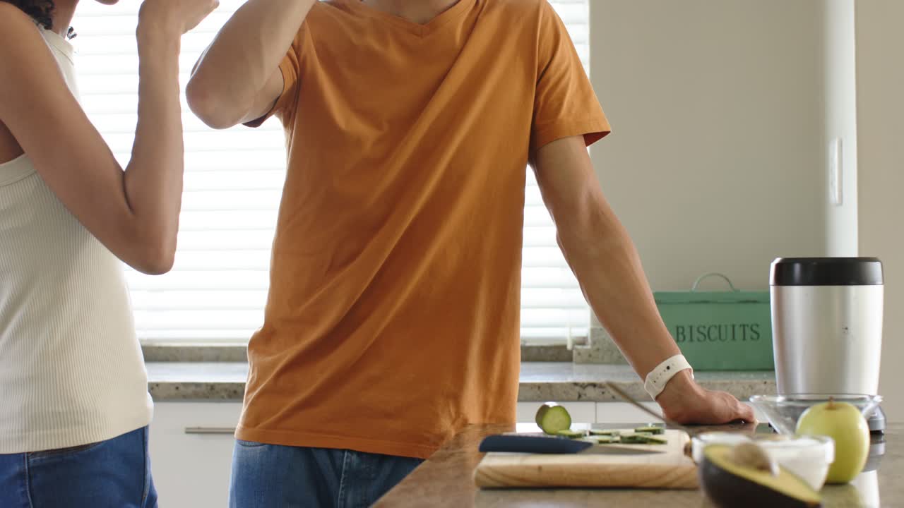 Coppia eterogenea che ride e assaggia frullati verdi al bancone della cucina di casa dopo aver frullato l'avocado