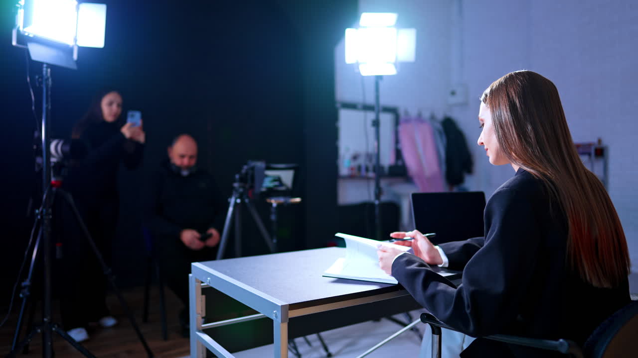 Naispuolinen juontaja mustassa bleiserissä työpöydän ääressä videotuotannon aikana. Nainen istuu työpöydän ääressä kannettavan tietokoneen kanssa studiossa, taustalla kuvausryhmä ja ammattimainen valaistuslaitteisto.