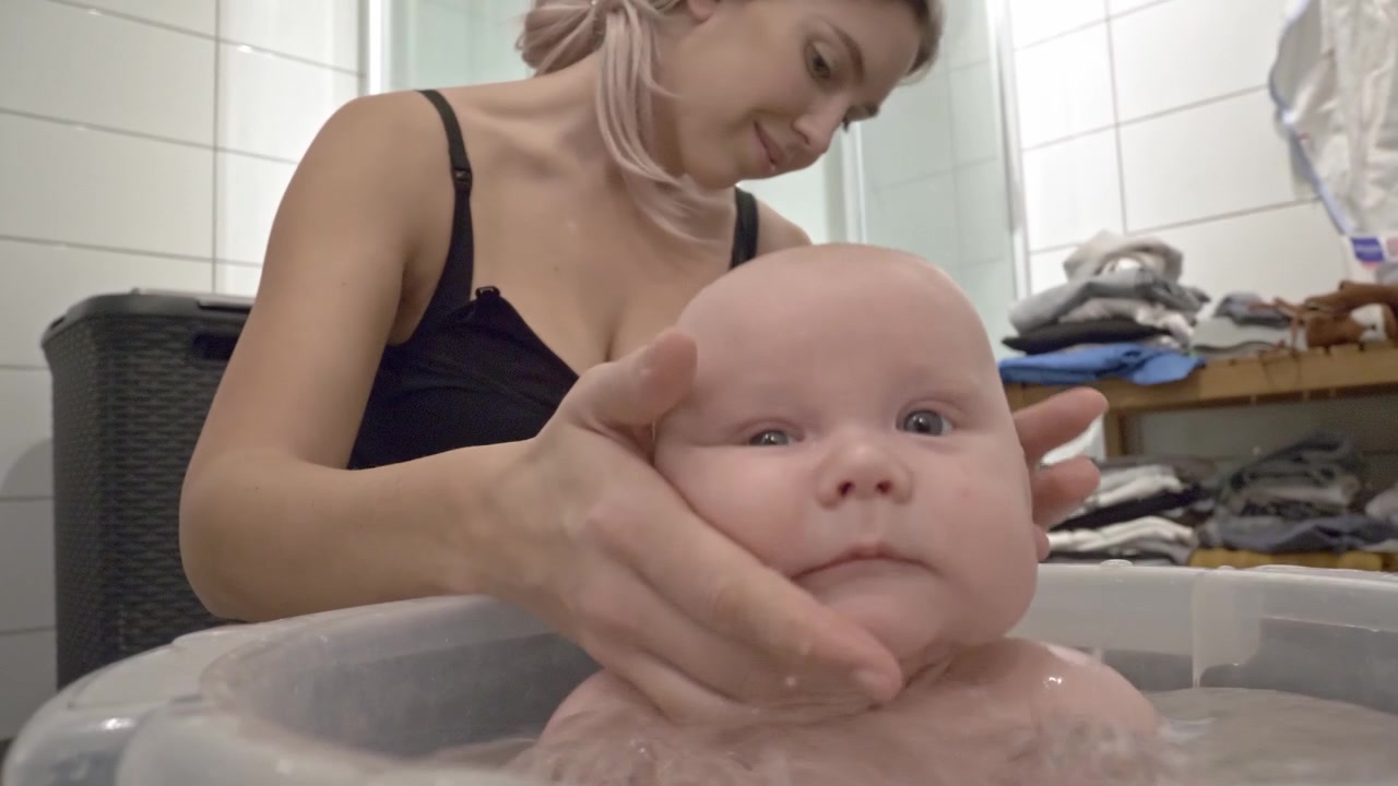 Bebê feliz espirrando na banheira com a mãe sentada no banheiro, segurando a cabeça do bebê