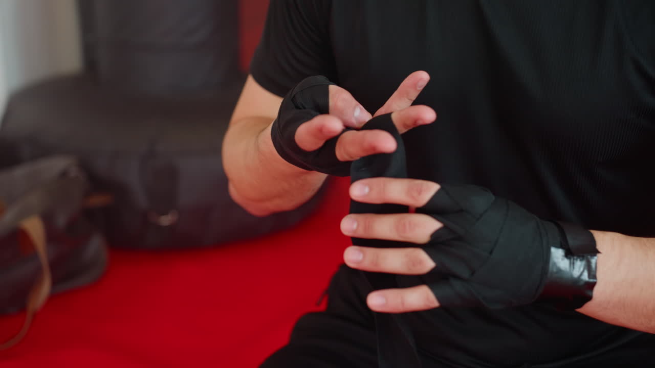 Boxer in black shirt prepares hands with black wraps before training, close up of focused wrapping technique, highlighting precision, discipline, strength, and readiness for combat inside gym with red mat