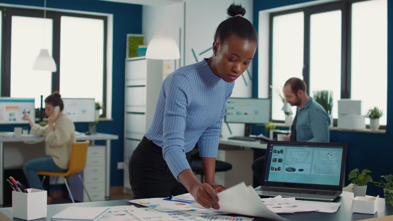 Busy african american in startup putting clipboard on desk looking at papers with sales charts comparing results