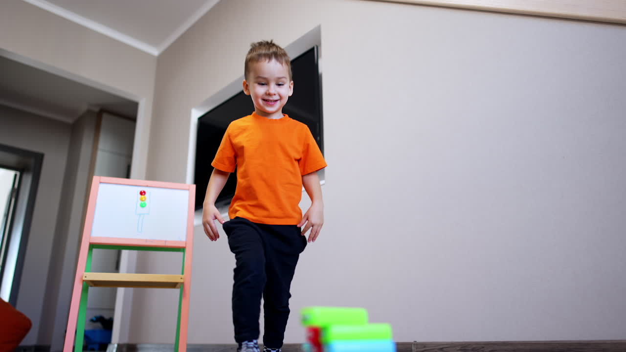 Smiling kid wearing orange t-shirt plays with bricks. Low angle view at the toddler building a tower of blocks.