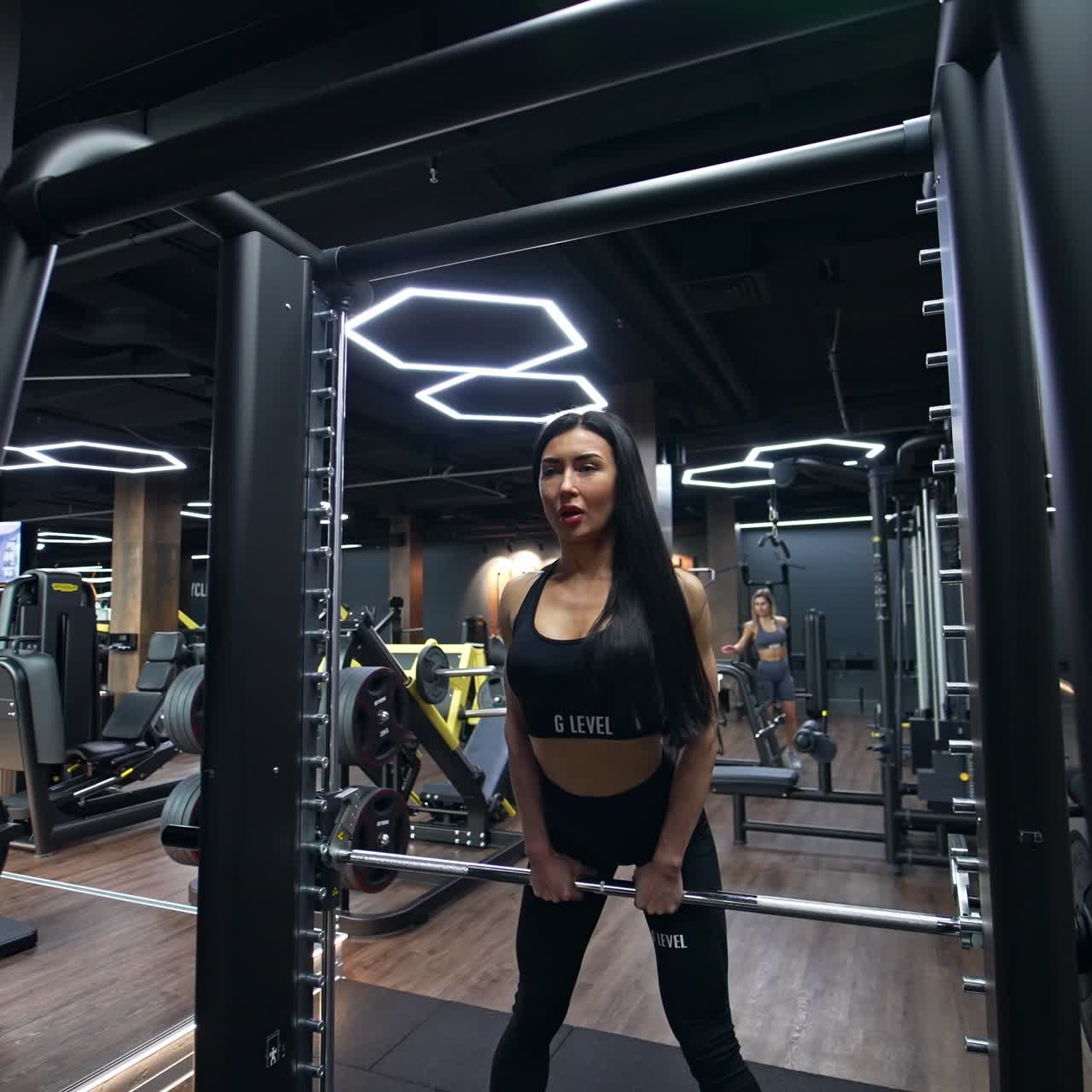 Una morena atractiva, en forma y con pelo largo pasa por delante del gimnasio. Una mujer se acerca a un soporte y levanta una barra horizontal para hacer ejercicio.