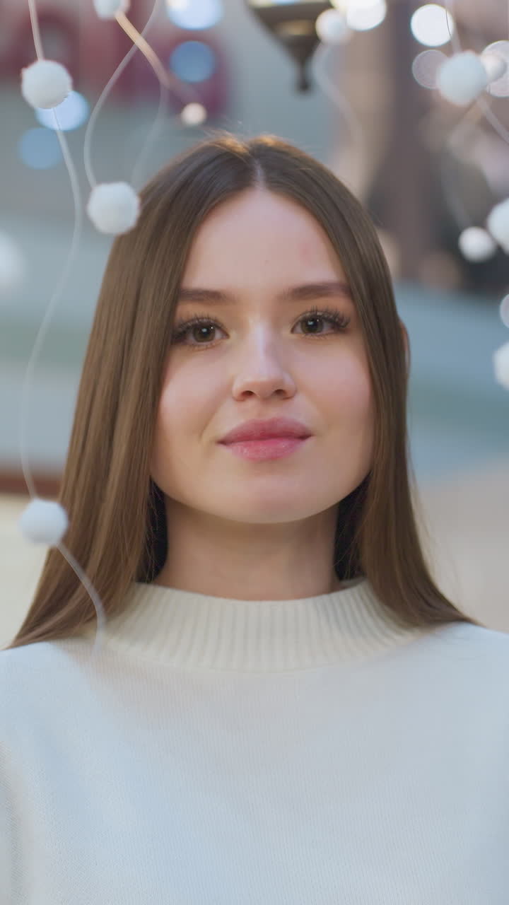 Beautiful lady with an adorably warm smile poses near a Christmas tree, delicately interacting with a hanging ornament, capturing a festive moment in a mall