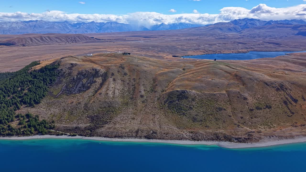 Sideway drone shot showing Lake Tekapo’s turquoise water, Mount John hill, and distant alpine mountains